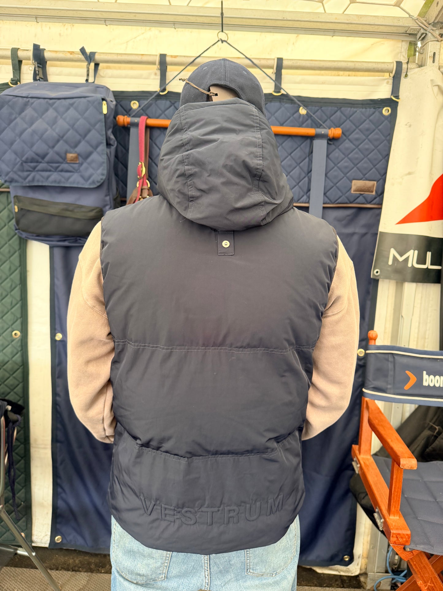 A person viewed from behind wearing a dark navy puffer vest with a hood and light beige sleeves underneath, standing in front of a display of bags and gear inside a tent.