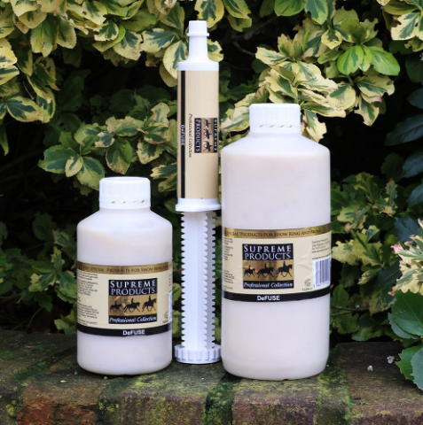 Three white plastic bottles and a white syringe-style dispenser with beige labels reading Supreme Products, placed outdoors on a brick surface in front of green variegated foliage.