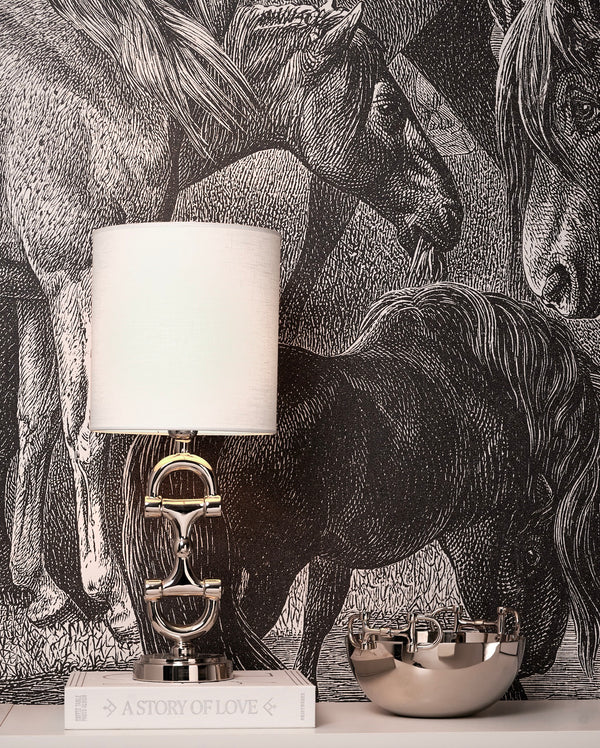 Silver bit lampstand with white lampshade stood on a book with a dark horse wallpaper background
