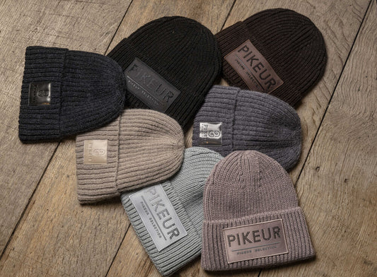 Six ribbed knit beanie hats in different muted colors are arranged on a rustic wooden surface. Each hat has a rectangular leather patch on the front with the brand name 'PIKEUR' printed on it.