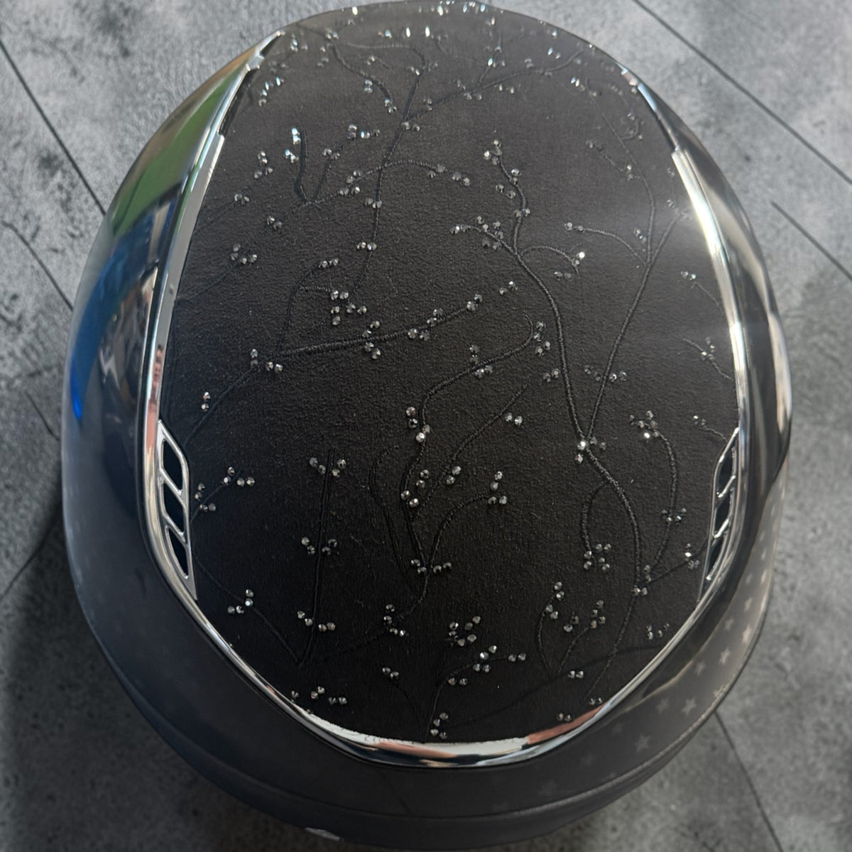 A glossy black horse riding helmet with a delicate silver rhinestone floral pattern on the front, resting on a gray tiled floor.