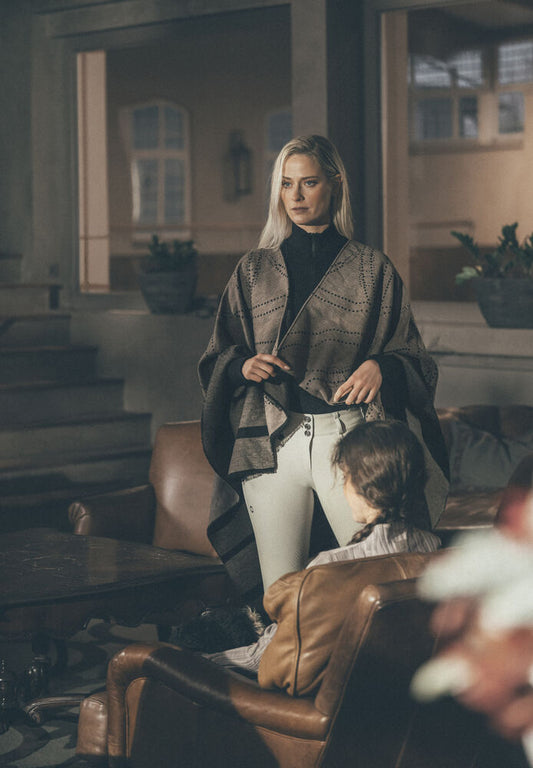 A woman stands in a dimly lit indoor room wearing a patterned brown shawl over a black top and white pants, holding a black handbag. A child is seated in a brown leather armchair in the foreground.