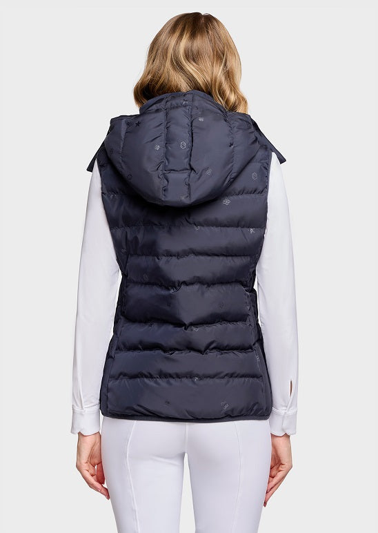 A woman shown from the back wearing a dark navy hooded puffer vest over a white long-sleeve top and white pants, against a light gray studio background.