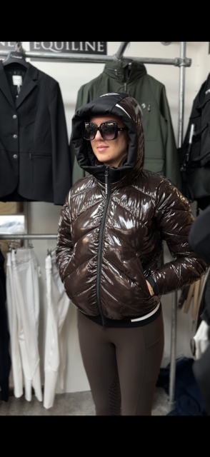 A woman wearing a shiny dark brown puffer jacket with a black zipper and black hood, styled with black sunglasses and brown leggings, standing in a clothing store.