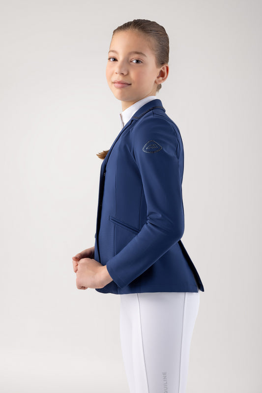 Young girl wearing a blue equestrian jacket and white pants on a plain background
