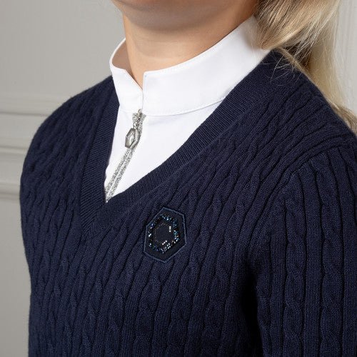 Close-up of a person wearing a navy blue cable-knit V-neck sweater over a white collared shirt, with a silver necklace and a small hexagonal embellished chest detail visible.