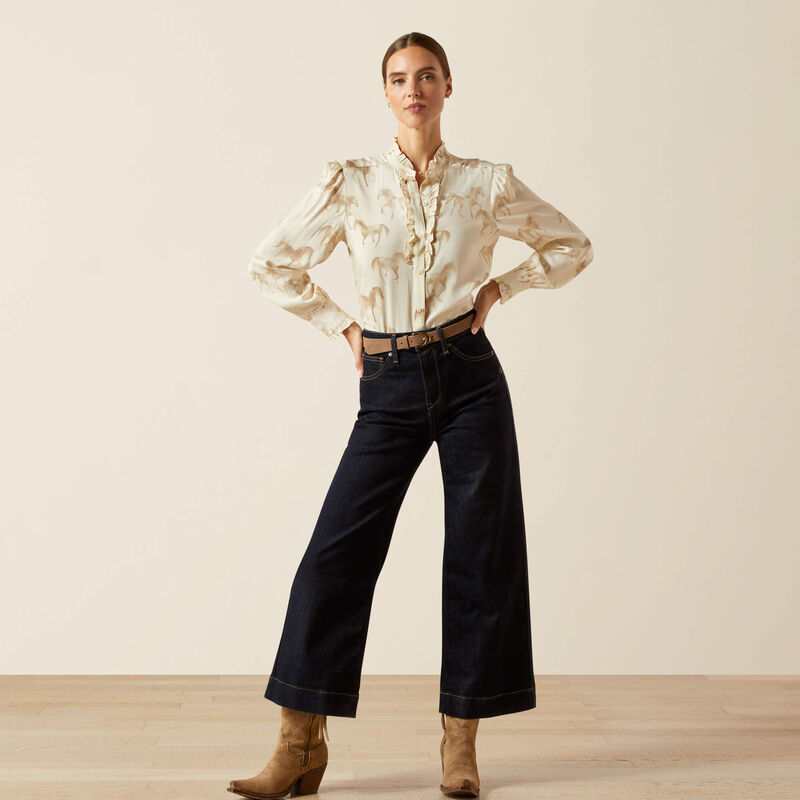 Woman wearing a horse print blouse, dark jeans, and brown boots against a plain background