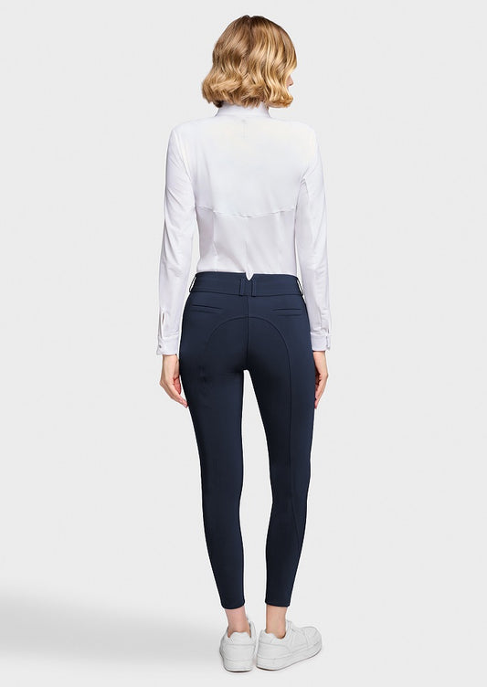 A woman viewed from the back wearing a white long-sleeve blouse, navy blue fitted pants, and white sneakers against a light gray studio background.