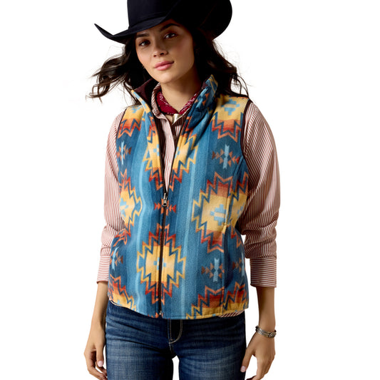 A woman wearing a sleeveless zip-front vest with a colorful geometric Southwestern pattern in blue, yellow, orange, and red over a striped shirt, paired with a black cowboy hat and jeans, against a plain white background.