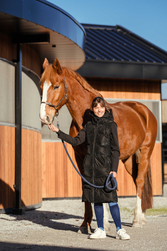 Woman in a long black sparkly vest holding a chestnut horse in front of a barn