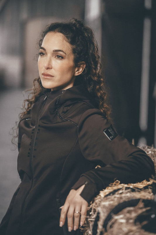 A woman with curly brown hair wearing a dark brown technical jacket, leaning against a textured hay bale in a dimly lit, industrial setting.