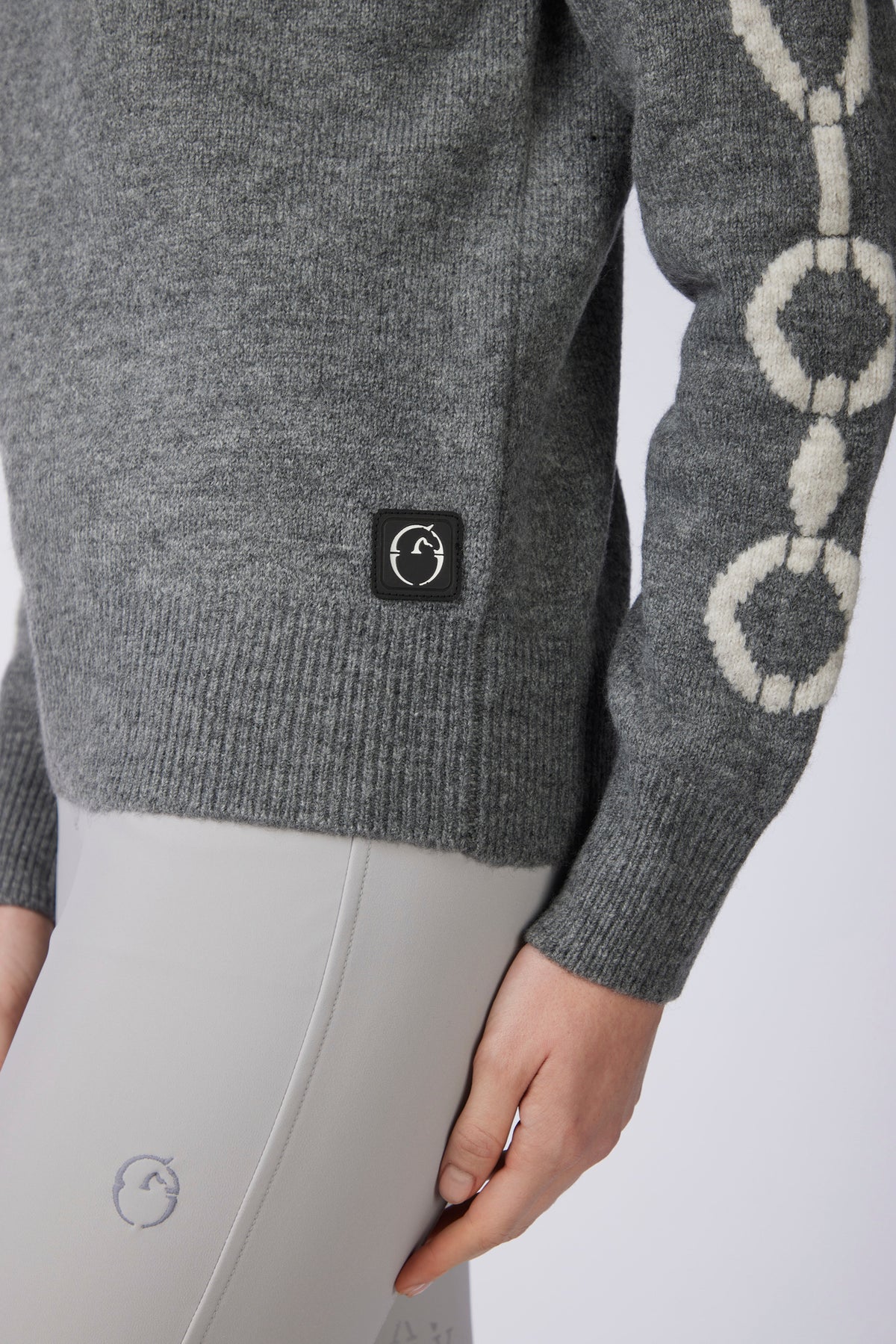Close-up of a person wearing a heather gray knit sweater with a black square logo patch on the lower left chest and a cream circular knit pattern on the sleeve, paired with light gray leggings.