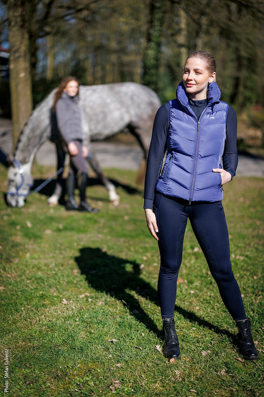 Woman wearing a blue gilet standing in front of a blurred gray horse