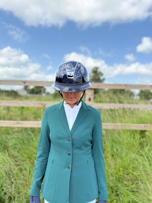 Person wearing a teal equestrian jacket and helmet in an outdoor setting with grass and a wooden fence.