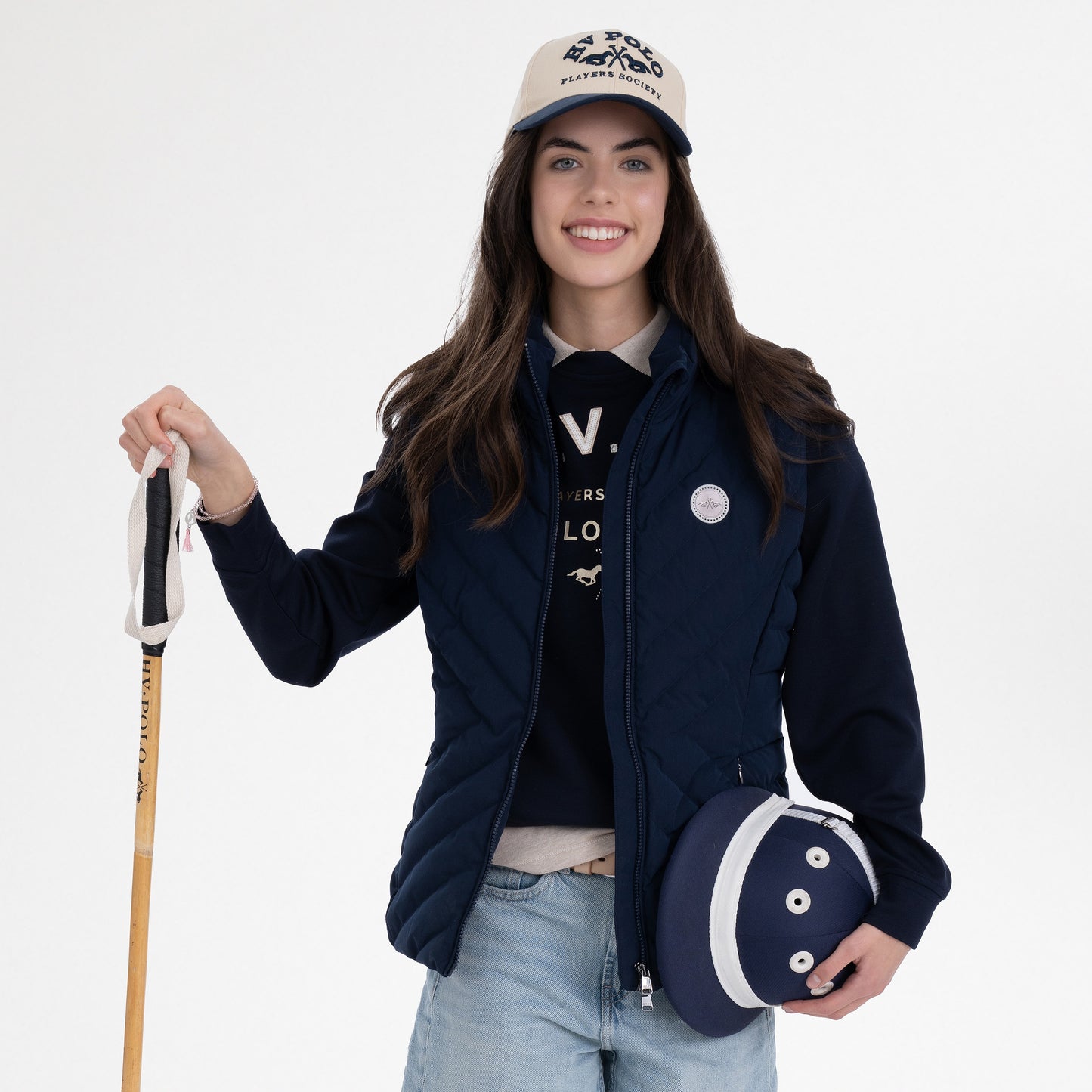 Girl wearing a navy gilet, navy hoodie, navy and beige cap and blue jeans, holding a polo whip and a polo hat on a white background