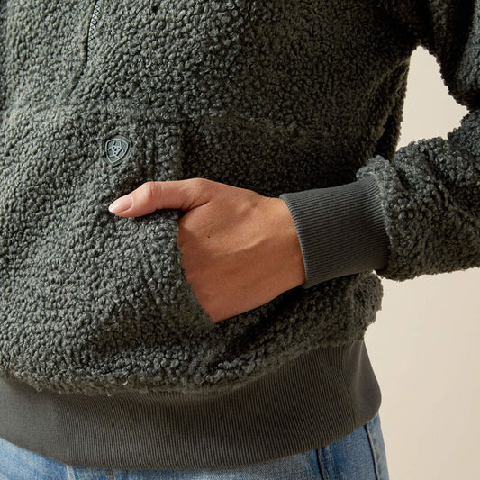 Person wearing a dark green sherpa jacket with a hand in the pocket against a neutral background