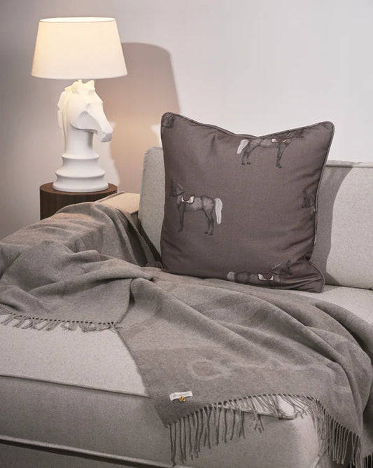 Gray pillow and throw blanket on a sofa with a lamp and decorative horse head sculpture in the background.