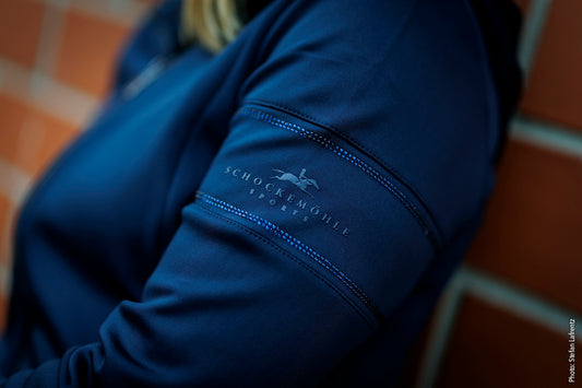 A close-up shot of a person wearing a dark blue athletic jacket, focusing on the upper sleeve which features the Schockemohle Sports logo and name in white, set against a blurred brick wall background.