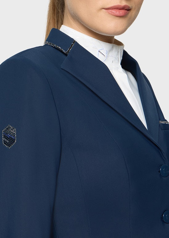 Close-up of a navy blue tailored blazer worn by a model, featuring decorative gold embroidered trim along the collar and a small embroidered crest patch on the sleeve, paired with a white collared shirt underneath.
