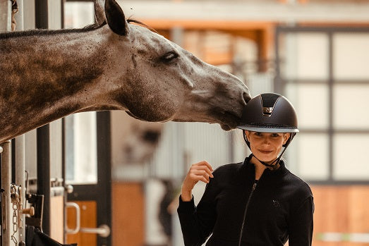 Girl wearing a Samshield helmet and a grey horse nuzzling her in a barn setting