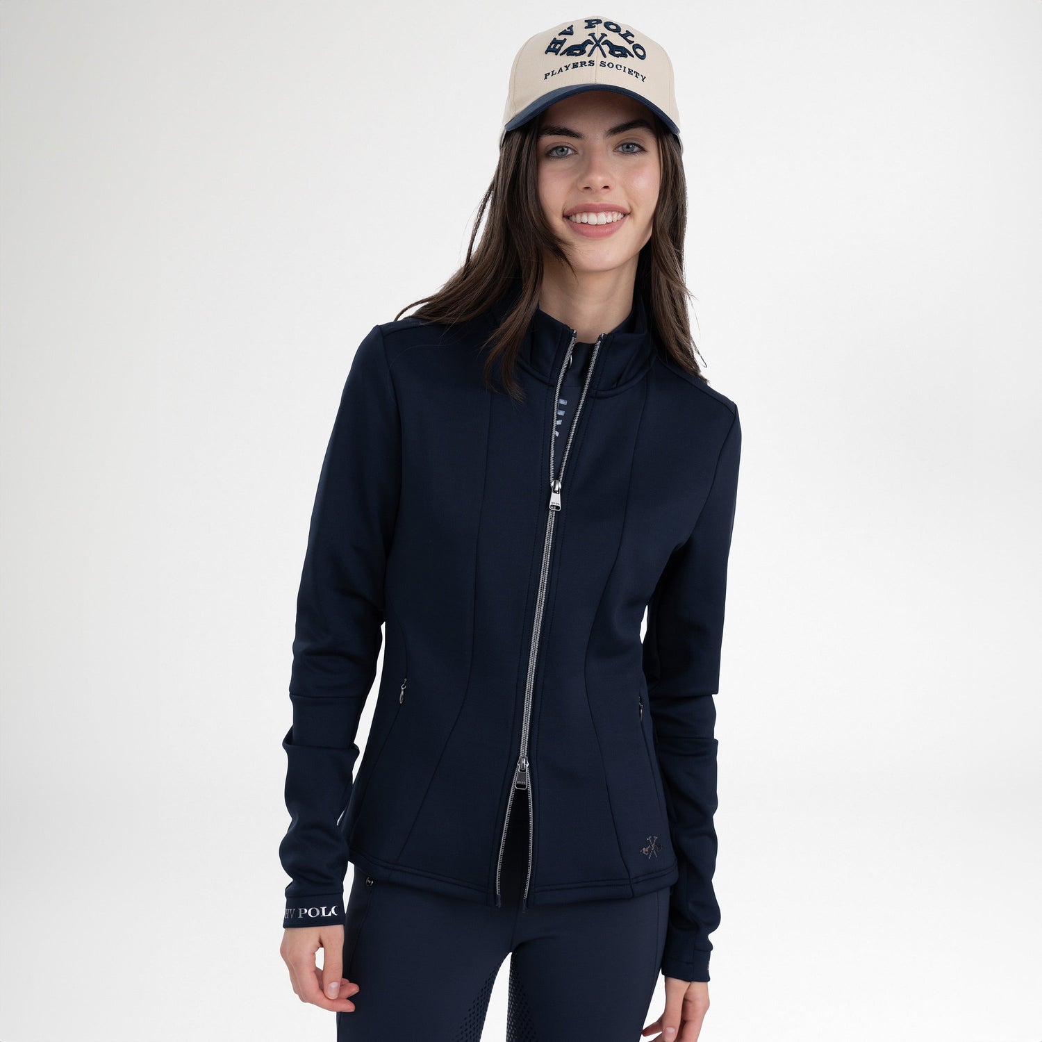 Girl wearing a navy full zip jacket, a pair of navy tights and a navy and cream baseball hat on a grey background 