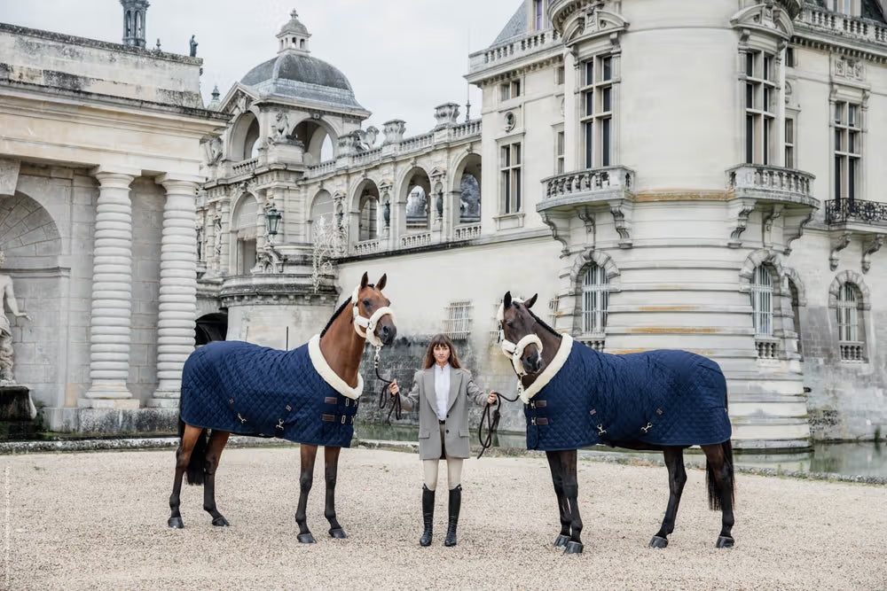 Two horses wearing navy Kentucky Horsewear rugs stand beside a person dressed in riding attire in front of an elegant historic stone building with arches and tower