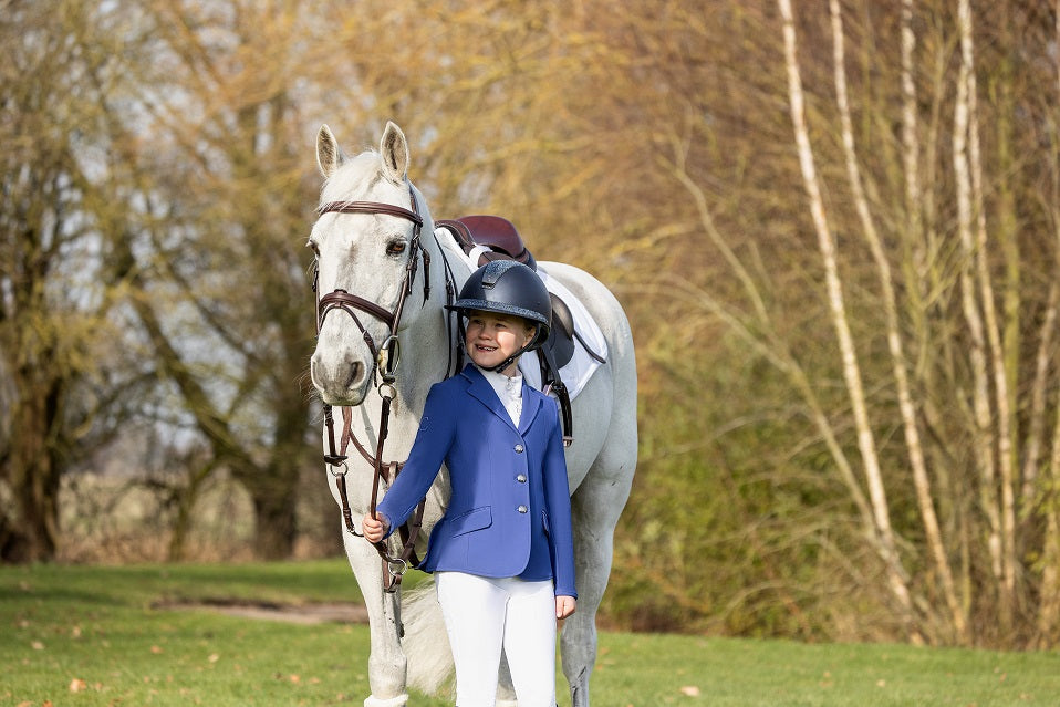 Young girl dressed in blue wearing a blue helmet holding a grey pony in an outdoor setting