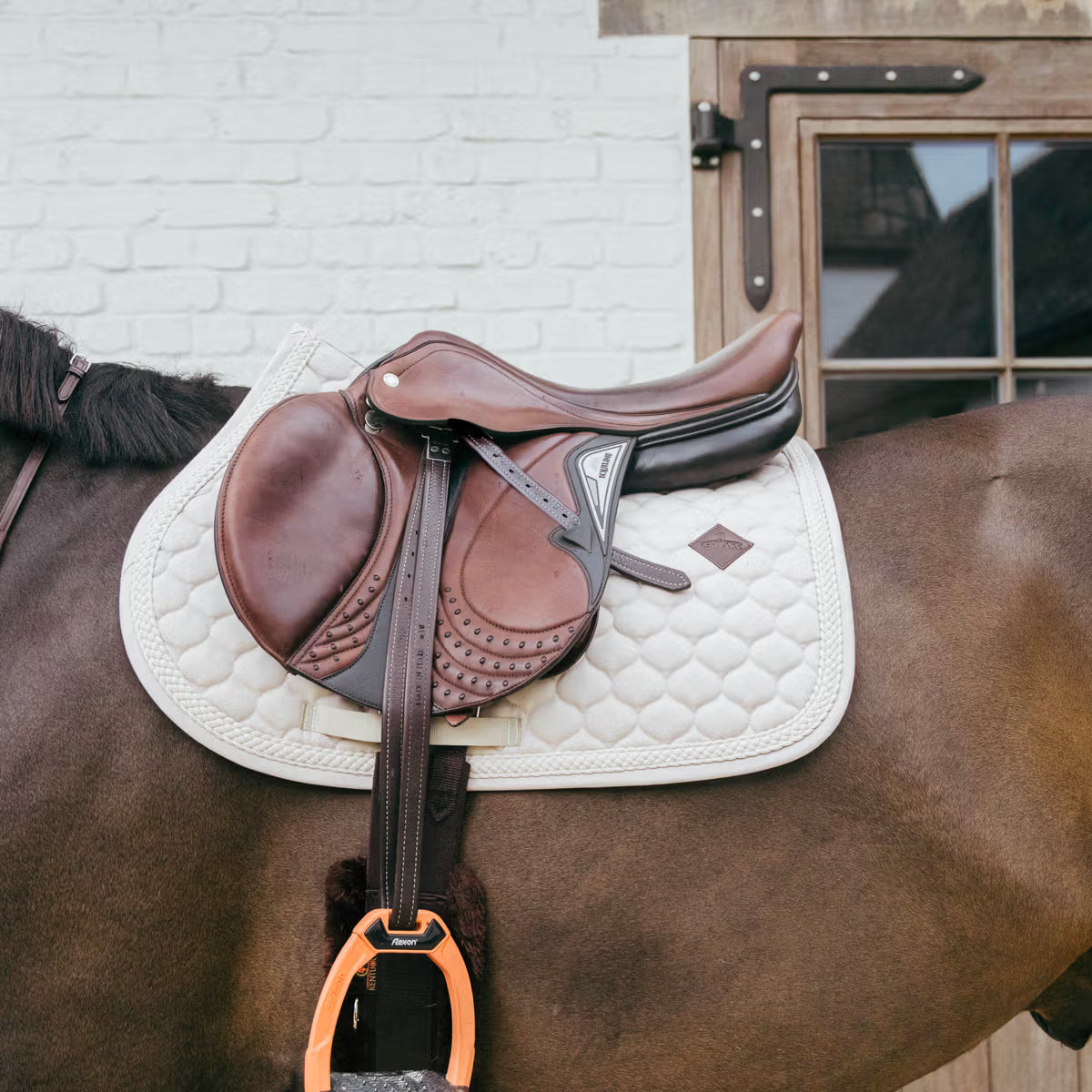 Beige saddle pad on a horse with a brown Equiline saddle and orange Flex On stirrups with a barn background