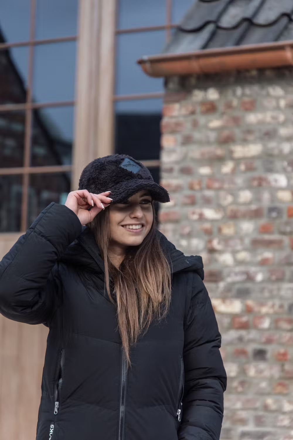 Person wearing a black Kentucky Rider puffer jacket and black faux-fur cap, smiling outdoors in front of a rustic brick building with large windows