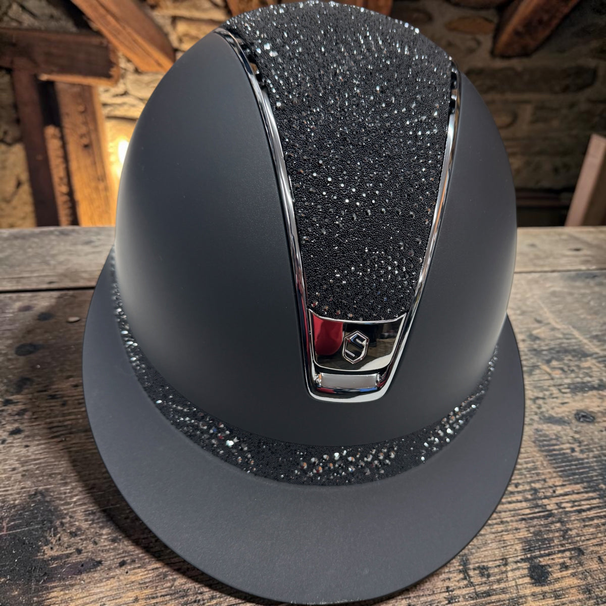 A matte black riding helmet with a glittery black crown insert, a silver chrome downpipe, and a matching glittery black brim, displayed on a rustic wooden table.