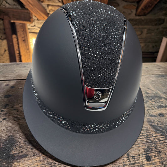 A matte black riding helmet with a glittery black crown insert, a silver chrome downpipe, and a matching glittery black brim, displayed on a rustic wooden table.