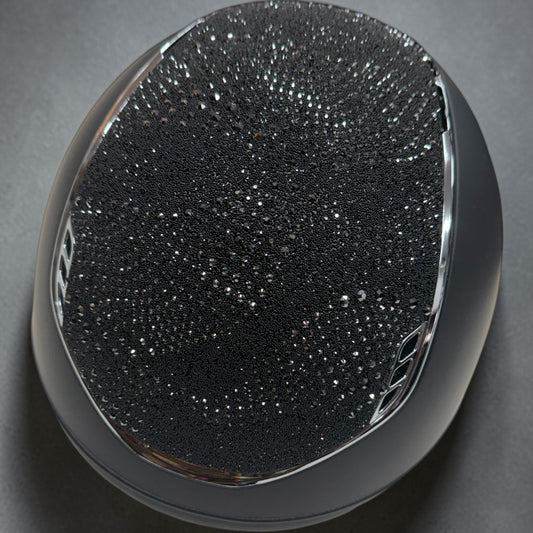 A close-up studio shot of a glossy black oval object with a textured, sparkly black surface and a polished metallic rim, set against a dark gray background.