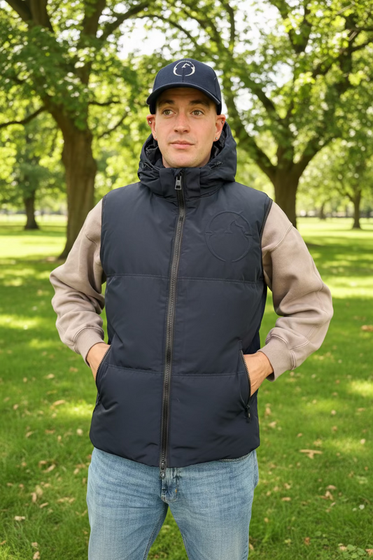 A man wearing a dark gray zip-up puffer vest over a beige and gray long-sleeve shirt, blue jeans, and a black cap, standing inside a tent-like stall with tack and chairs in the background.