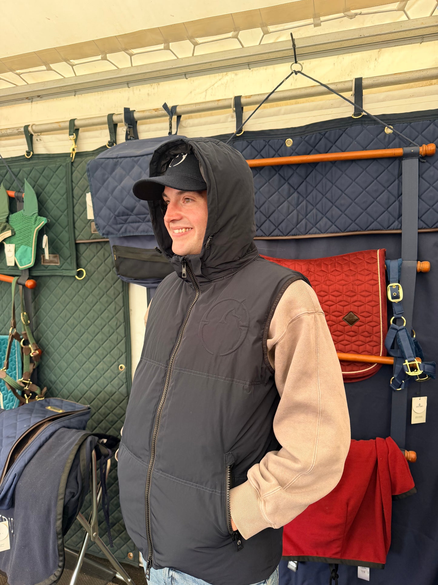 A man wearing a black quilted zip-up puffer vest over a beige hoodie and a black cap stands in front of a display of horse tack and saddles inside a stable or tack room.