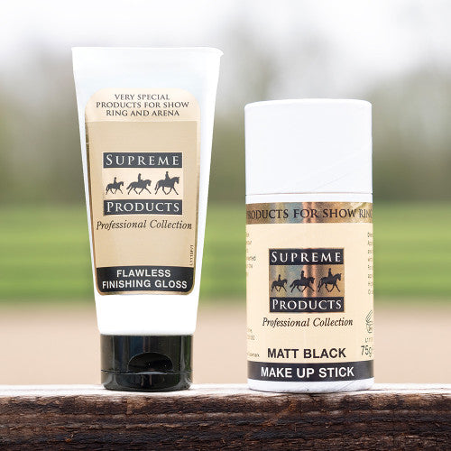 A white squeeze tube of Supreme Products Flawless Finishing Gloss and a white stick container of Supreme Products Matt Black Make Up Stick, both standing on a rustic wooden surface with a blurred outdoor background.