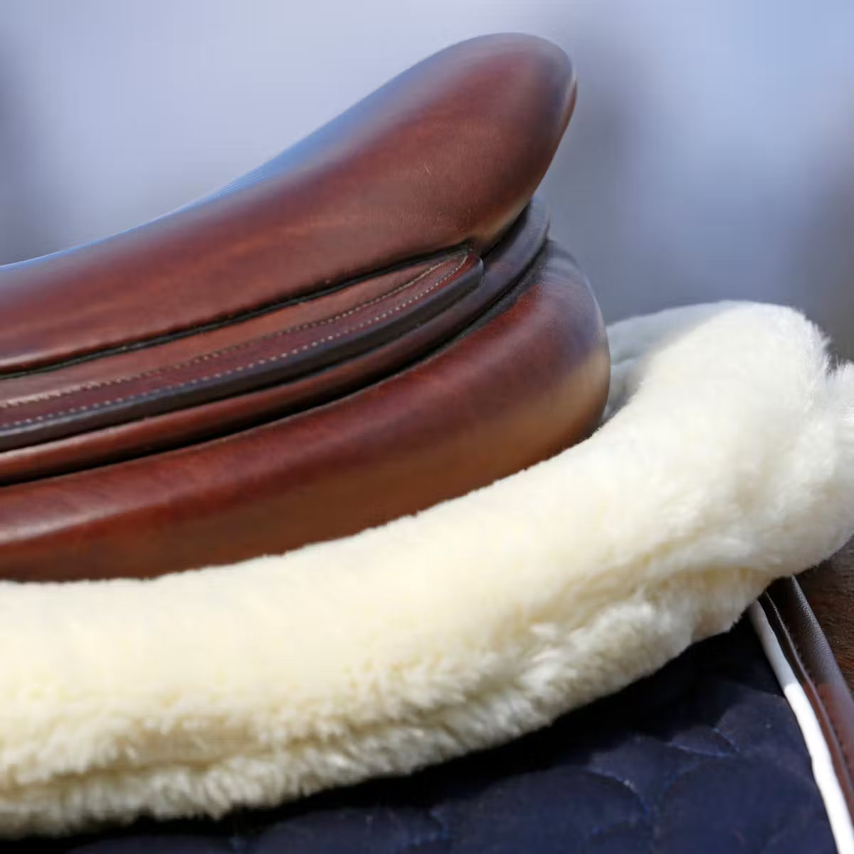 Close-up of a brown leather saddle with a white sheepskin pad on a blurred background