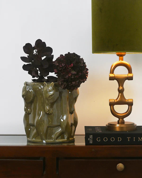 Decorative vase with horse design, black flowers, and a gold lamp on a wooden surface.