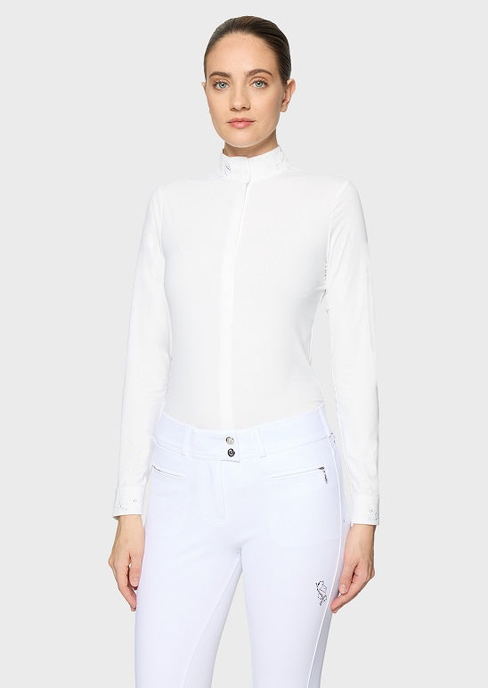 A woman modeling a white long-sleeve mock neck equestrian shirt and white riding breeches against a light gray studio background.