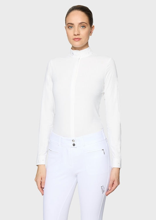 A woman modeling a white long-sleeve mock neck equestrian shirt and white riding breeches against a light gray studio background.