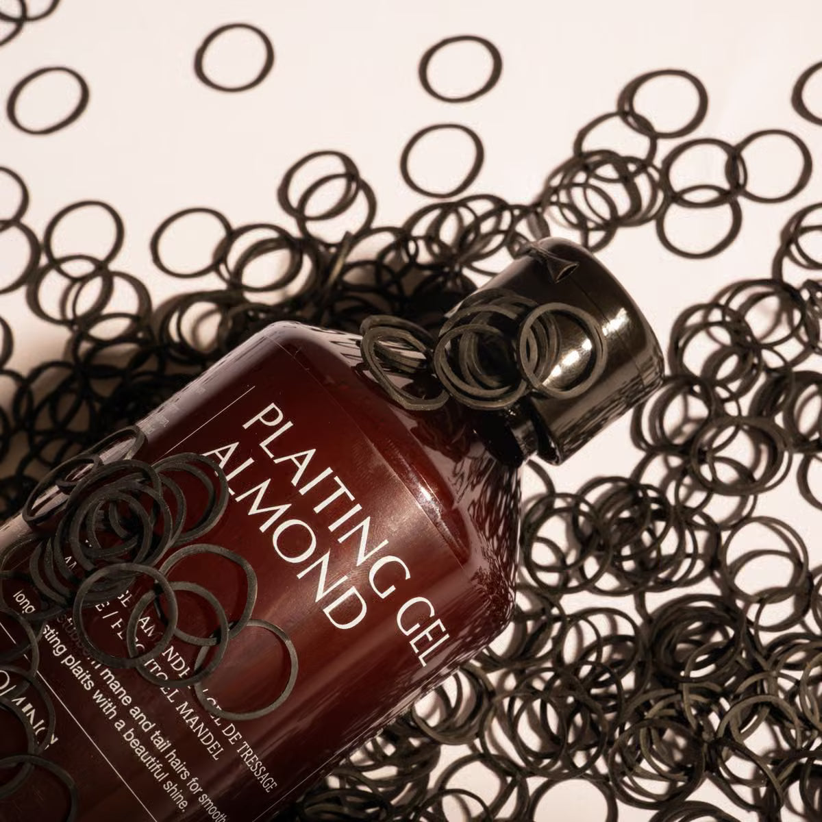 A bottle of Plaiting Gel Almond lies on a light background surrounded by scattered black hair rings.