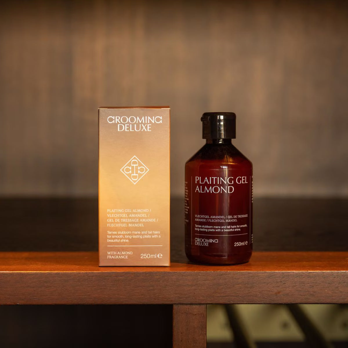 A bottle of Plaiting Gel Almond grooming product next to its packaging box, both in a warm amber-brown tone, displayed on a wooden shelf against a dark wood background.