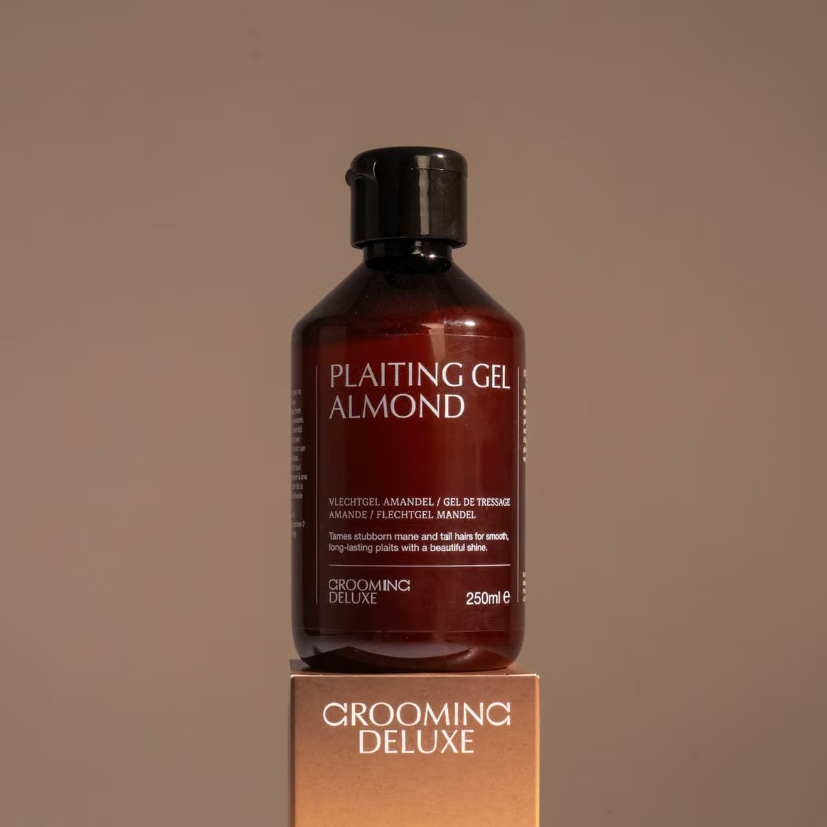 A bottle of Plaiting Gel Almond grooming product from Grooming Deluxe, displayed on top of its metallic gold box against a solid taupe background.