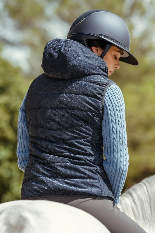 Person wearing a blue quilted jacket and helmet, sitting on a horse with a blurred natural background