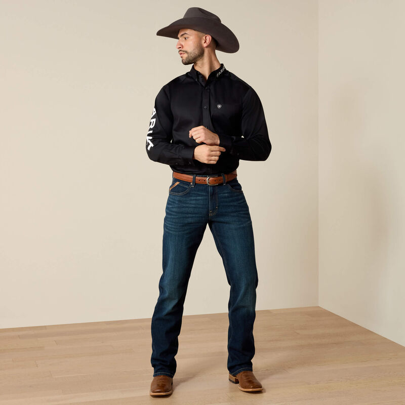 Man wearing a black shirt, blue jeans, and a cowboy hat in a room with beige walls and wooden floor.