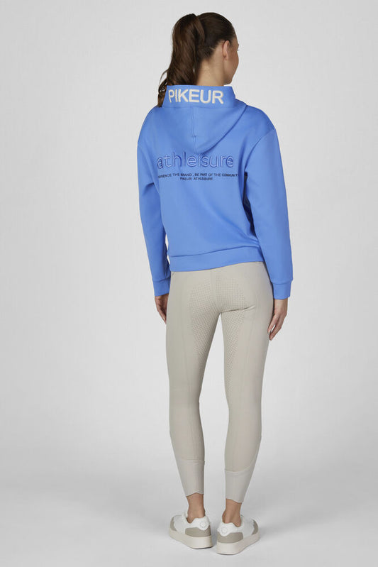 A woman is shown from the back wearing a bright blue athletic hoodie with 'PIKEUR' printed on the back collar and 'athleisure' on the upper back, paired with light beige leggings and white sneakers, against a light gray studio background.