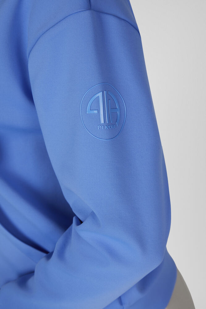 A close-up of a blue long-sleeve top or sweatshirt showing the upper sleeve with a tonal circular logo patch featuring the brand Pikeur.