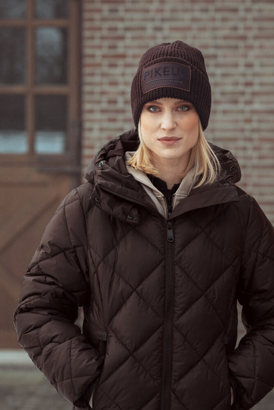 A woman wearing a dark brown diamond-quilted puffer jacket and a matching dark brown knit beanie with a visible 'PIKEU' brand patch, standing in front of a blurred brick building.