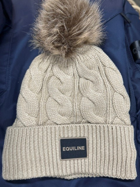 A close-up of a cream-colored cable-knit beanie with a brown faux fur pom-pom, laid flat on a navy blue jacket. The beanie features a black brand patch reading 'EQUILINE'.