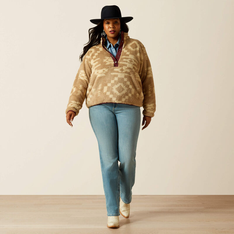 Person wearing a beige patterned sweater, blue jeans, and a black hat on a plain background