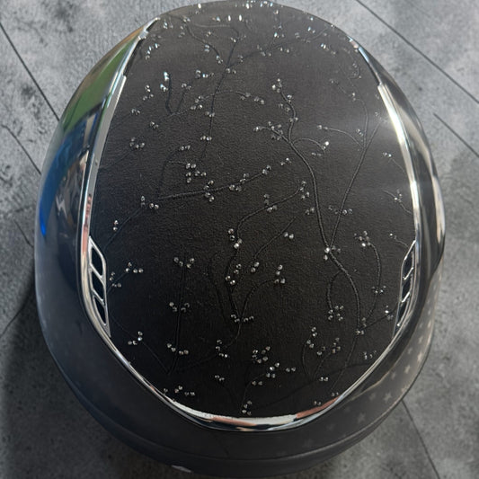 A glossy black horse riding helmet with a delicate silver rhinestone floral pattern on the front, resting on a gray tiled floor.